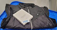 Black men's medium heated vest folded with visible heating control button and original packaging.
