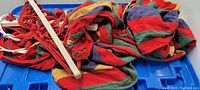Photo of a multicolored cloth hammock folded showing red, green, yellow, purple stripes with red rope ends and metal hanging rings on blue plastic background.