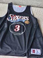 Front view of black mesh Allen Iverson jersey with Philadelphia 76ers logo and number 3, showing tag details.