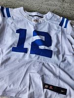 Front view of white football jersey with blue number 12, blue stripes on shoulder, showing mesh and fabric material.