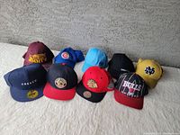 Photo of 9 assorted baseball and sports caps on light surface against textured gray wall