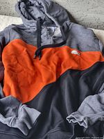 Front view of Lacoste warm-up hoodie showing gray, orange, and black color blocks with drawstring hood.