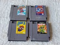 Four Nintendo NES game cartridges arranged on a white surface showing front labels: Super Mario Bros. 2, Mega Man 2, Super Mario Bros. 3, and Teenage Mutant Ninja Turtles: The Arcade Game.