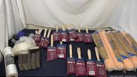 Full lot showing paint brushes, sanding blocks, roller covers, respirator mask, hand-masking plastic, and masking string on dark cloth backdrop.