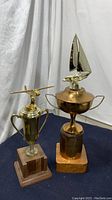 Two vintage trophies displayed on dark fabric backdrop, one with a sailboat figure on metal cup with wooden base, the other with airplane figure on tall metal cup with wooden base.