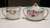Front view of Nippon creamer and sugar bowl with lid, showing detailed hand painted pink roses and green leaves with pale blue background and gold trim.