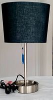 Full view of lamp showing black fabric oval lampshade, silver-toned stick lamp body and circular metallic base with power outlet and cord.