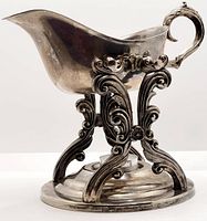 Full profile side view of silver plated gravy boat resting on ornate warming stand with intricate scroll design and curving handle.