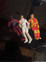 Three loose GI Joe action figures displayed together on a dark surface. One vintage figure in white, one in red and black suit, and one in yellow and red suit.