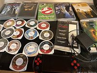 Overview of Sony PSP handheld system, multiple UMD game discs, some in cases, charger with missing part