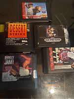 Photo of five Sega Genesis game cartridges and cases laid out on a glass surface, showing the titles Desert Strike, Barkley Shut Up and Jam!, JX3 Arena, an unlabeled cartridge, and a baseball game.
