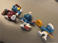 Photo of three vintage Smurfs figurines including a baker, graduation, and shovel Smurf laid out on grey surface