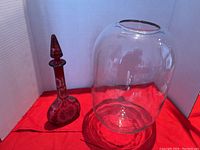 Cranberry etched glass decanter with stopper and large clear glass vase side by side on red cloth inside white backdrop box.