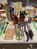 Overview of kitchen utensils and items on table including knives, scissors, ladle, garlic press, pineapple container, and other kitchen tools.
