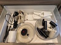 Open drawer showing various kitchen implements including Black & Decker hand mixer, salad spinner, mandolin parts, juicer and metal tongs.