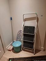 Photo showing the full lot including the 3-step ladder, garbage bin, ironing board top, and mop in a corner of a room with wood flooring and white walls.