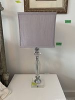 Full view of one lamp showing purple square fabric shade on crystal and metal base on white surface.