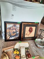 Two framed prints on floor; musical instruments print in black frame with gold inner border; floral print in large ornate brown frame