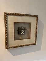 Framed shadow box with ornate metal piece centered, gold-toned carved frame, beige textured background matting.