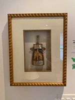 Frontal view of shadow box frame containing a decorative metal flask with wood inlays, mounted on light textured backing inside a gold-tone frame.