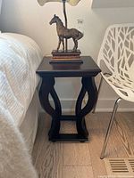 Side table positioned between bed and a white chair on hardwood floor; horse statue lamp on top not included