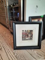 Framed signed print of a street cafe scene leaning against a wall on a wooden floor.