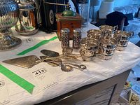 Photo showing six silver plate cup holders, three serving utensils including cake server, and pair of salt and pepper shakers on table.