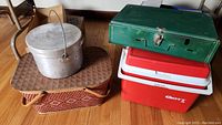 Overview photo showing all items including Coleman green camping stove, red and white Gott cooler, aluminum cooking pot, and woven picnic basket with wooden lid.