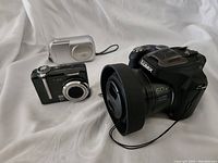 Three digital cameras - Panasonic Lumix DMC-FZ70 black with large zoom lens, Olympus silver compact camera, Kodak black compact camera