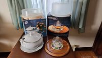 Photo showing ceramic soup tureen with lid and base plate, traveling cake and dip carriers on top of cardboard boxes, wooden cheese board with glass dome cover in front