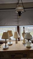 Five assorted lamps on cardboard box near window including hanging lamp, two milk glass beaded lamps with brass and milk glass bases, and ceramic lamps with shades