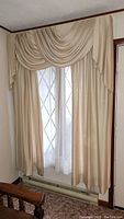 Beige woven curtain with swag and sheer panel on window next to wood chair and carpeted floor.