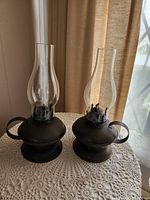 Two bronze oil lamps with glass chimneys, standing side by side on a white crochet tablecloth, showing complete assemblies from front angle.