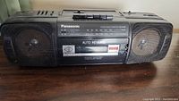 Front view of black Panasonic stereo radio cassette boom box with two speakers, cassette player, and buttons on top.