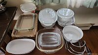 Photo shows an assortment of white CorningWare casserole dishes with lids, Pyrex rectangular glass baking dishes, white ceramic Anchor Hocking baking dishes, and metal racks all displayed on a table.