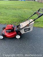Full side view of red Toro self propelled lawn mower with beige clippings bag on black handle, set on asphalt surface.