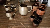 Overview of entire set on wooden surface showing metal pitcher, metal small goblet cups, wooden carved cups, and smooth wooden cups