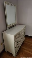 Photo of low white wooden dresser with multiple drawers and attached rectangular mirror against wall on hardwood floor, showing front and side.