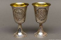 Front view of two silver Kiddush cups showing detailed Judaica scenes: one with Jerusalem cityscape, the other with the Western Wall.