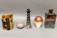 Five vintage pottery pieces arranged in a line: from left, a wavy-edged bowl with earth tones, a small pink and green bowl, a tall candleholder with black and white geometric patterns, a rounded beige-orange vase with black marbling, and a rectangular vase with geometric decoration.