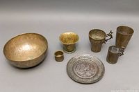 Seven old brassware pieces, including bowls, cups with decorative handles, and an etched plate, all with visible wear and patina.