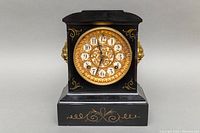Front view of black antique mantle clock with gold ornate dial, showing clock face and gold decorative accents.