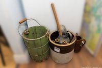 View of green painted wooden bucket with metal bands and a wooden handle next to a cream and brown stoneware crock filled with wooden and metal implements and a small copper/brass jug.