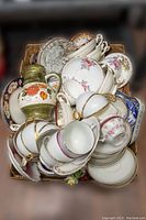 Top-down view of assorted teacups, saucers, crystal glasses, and a floral-painted ceramic pitcher packed in a box
