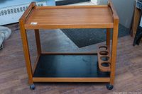 Front view of two-tier teak bar trolley on casters with top tray and black lower shelf featuring four circular bottle slots