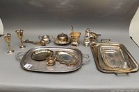 Full lot of silver plated servingware including trays, butter dish, gravy boat, goblets, sugar bowl, creamer, salad tongs, and small plates seen on a flat surface.