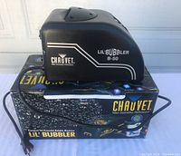 Front and side view of the Chauvet Lil Bubbler B-50 bubble machine placed on its original box. Shows the black body with brand and model identification.