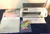 Front and left angle view of Cricut electronic cutting machine with accessories laid out including cutting mats and cutting tools in a plastic bag.