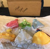 Five Disney princess-style miniature dollhouse dresses laid out on white tissue paper with cardboard box labeled 'Mini Dress Shoppe' in background.