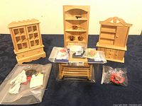 Three unfinished wooden dollhouse furniture pieces including a China cabinet, corner hutch with miniature pots, and smaller hutch with shelving and door. Miniature food items on table. Fabric clothes and wall decorations.